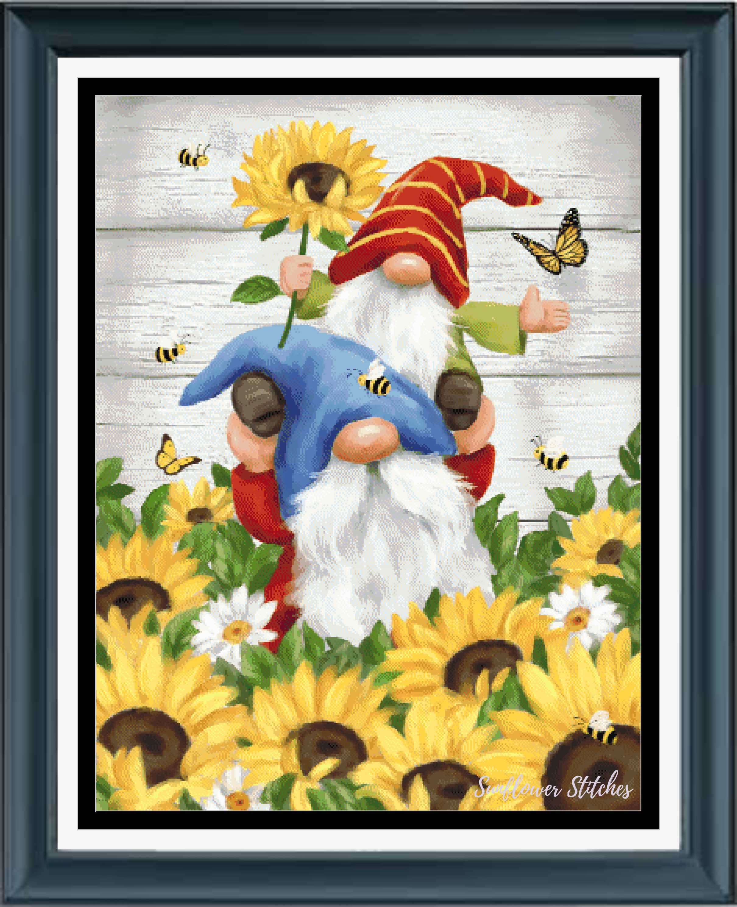 Framed decorative art print featuring two garden gnomes among yellow sunflowers and small bees, with a white wood-plank background and a dark blue frame.