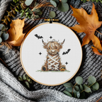 Finished mummy Highland cow Halloween counted cross-stitch pattern in an embroidery hoop, styled on a chunky knit blanket with autumn leaves and eucalyptus, by Sunflower Stitches.