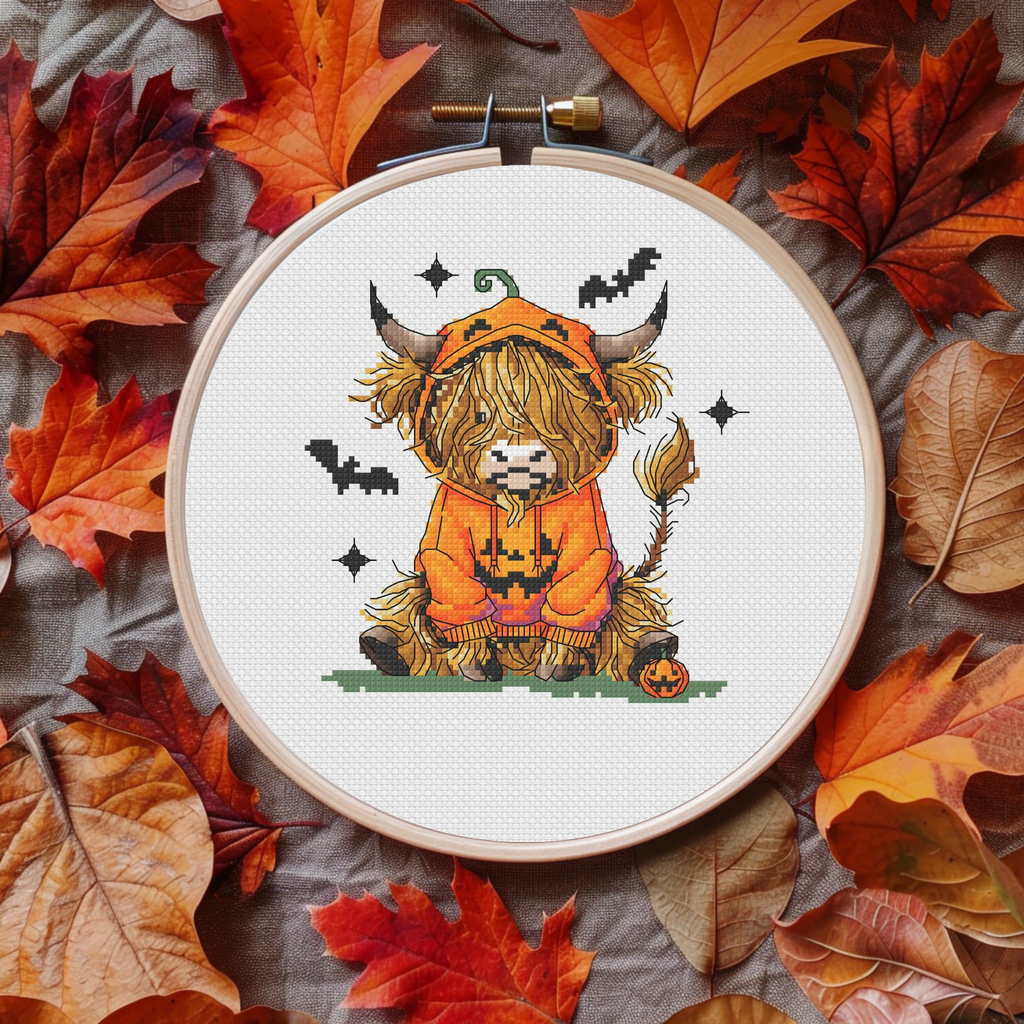 A cross-stitch embroidery hoop displaying a cute Highland cow in an orange pumpkin costume, surrounded by a flat-lay of colorful autumn leaves on a gray fabric background.