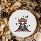 A circular embroidery hoop displays a cross-stitch design of a cute Highland cow dressed as a vampire, surrounded by autumn-themed flat-lay props like pinecones, dried leaves, and a candle.