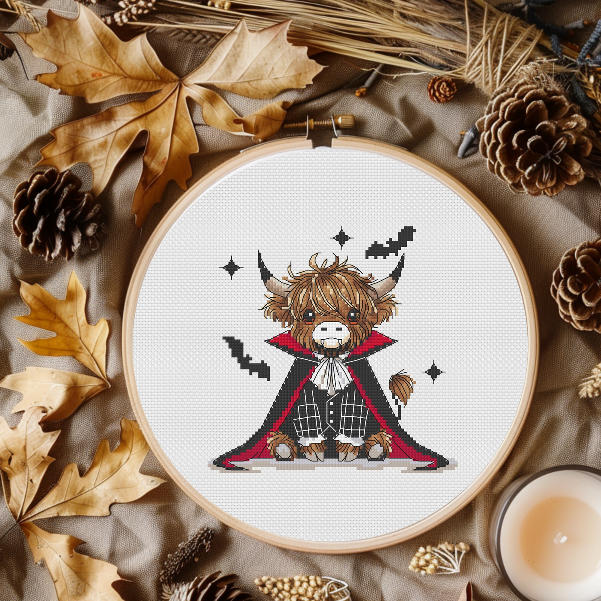 A circular embroidery hoop displays a cross-stitch design of a cute Highland cow dressed as a vampire, surrounded by autumn-themed flat-lay props like pinecones, dried leaves, and a candle.