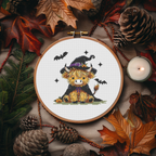 Finished witch Highland cow Halloween counted cross-stitch pattern in an embroidery hoop surrounded by autumn leaves, pinecones, and a lit candle, by Sunflower Stitches.