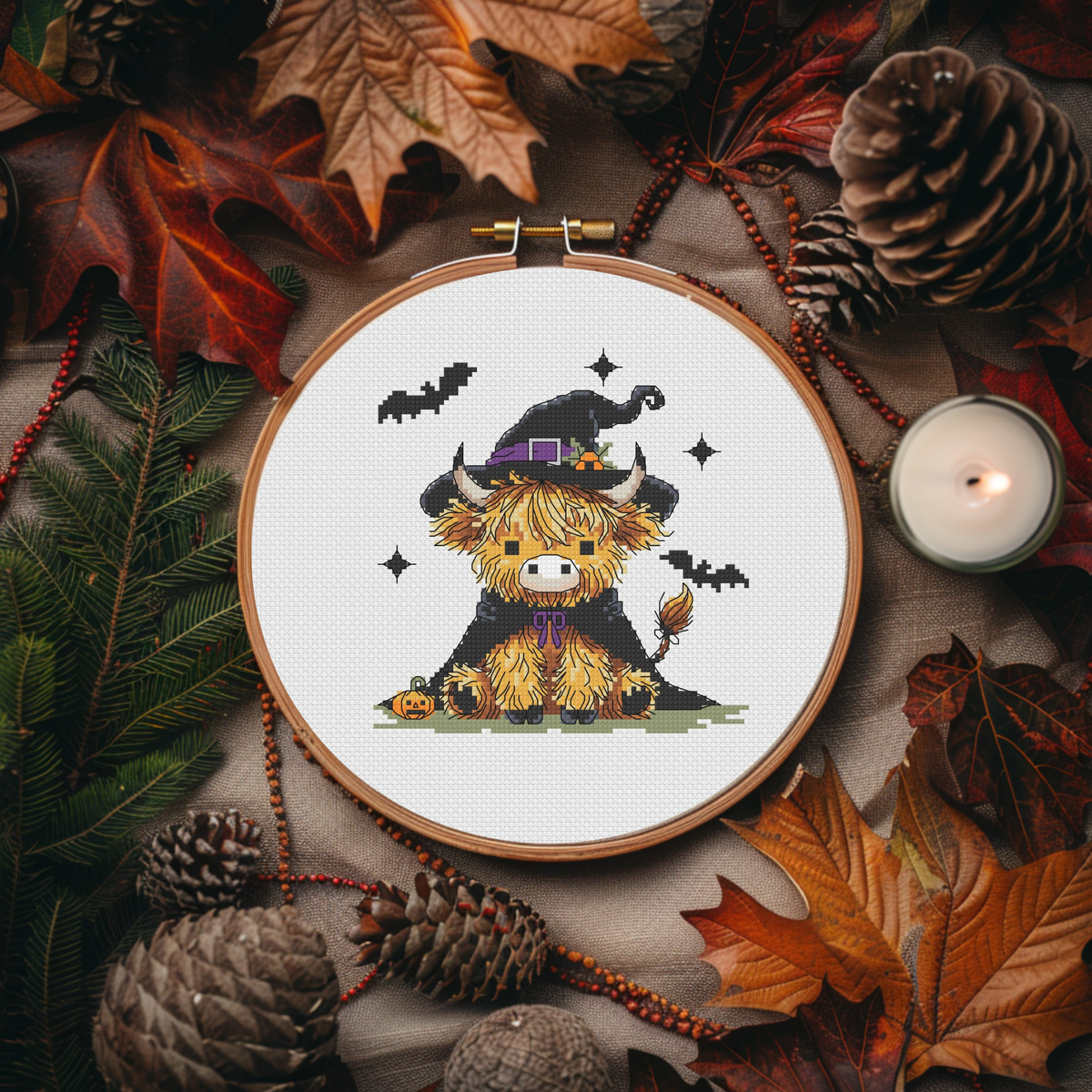 A circular embroidery hoop displays a cross-stitch design of a fluffy yellow Scottish highland cow wearing a black witch hat with a purple band. The hoop is surrounded by autumn leaves, pinecones, evergreen branches, and a lit candle.