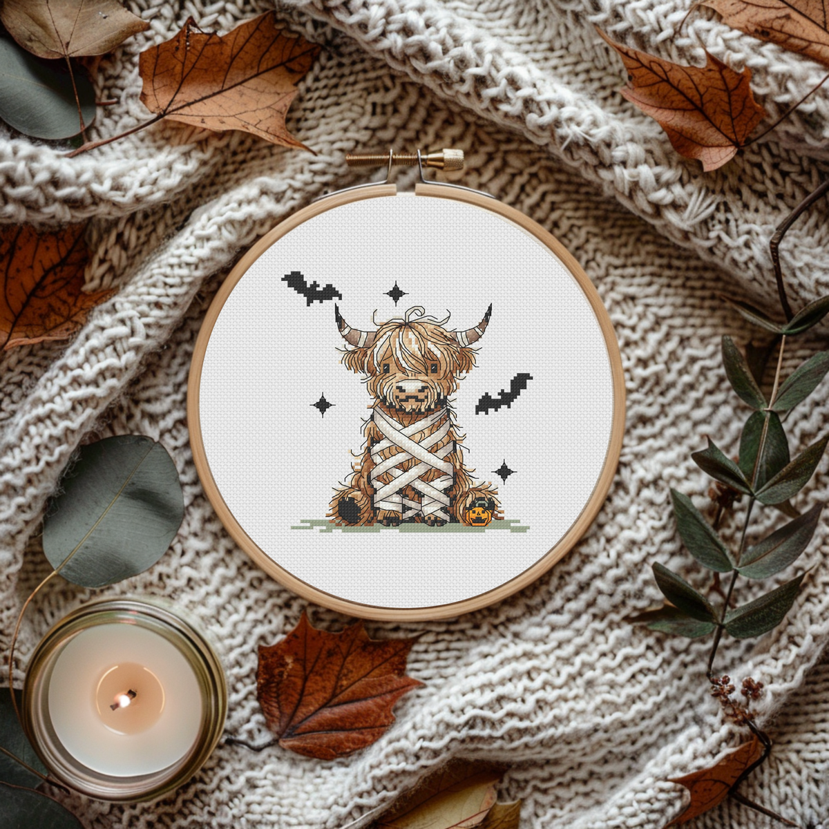 A circular embroidery hoop displays a finished cross-stitch design of a fluffy Highland cow dressed as a Halloween mummy, the hoop is surrounded by autumn leaves, a knitted blanket, and a lit candle.