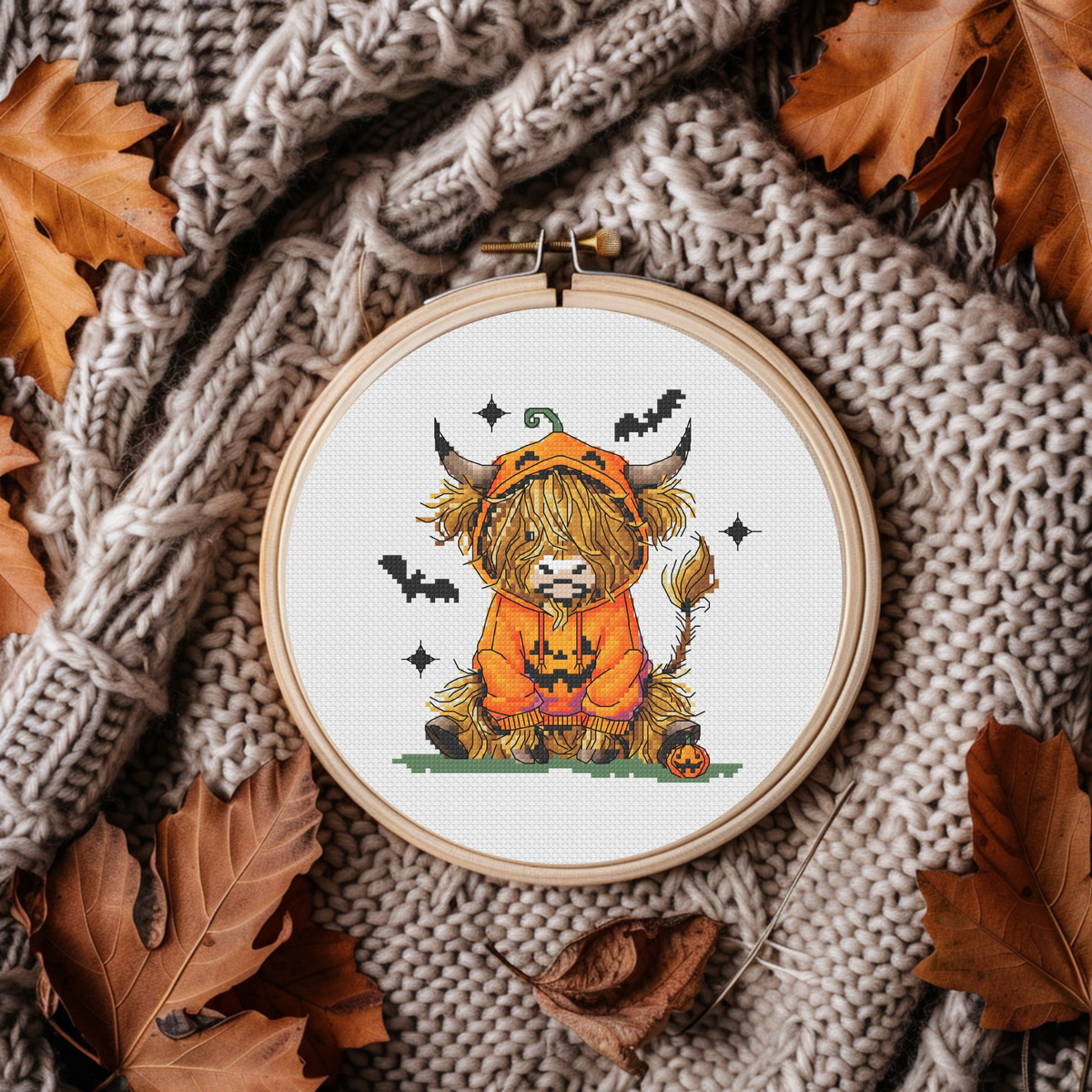 A circular embroidery hoop holding a cross-stitch design of a fluffy yellow highland cow in a pumpkin costume, surrounded by autumn leaves and a chunky knit beige blanket.