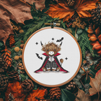 A circular embroidery hoop displays a finished cross-stitch design of a cute Highland cow dressed as a vampire, surrounded by a decorative autumn flat-lay of pinecones, acorns, and leaves.