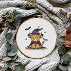 Finished witch Highland cow Halloween counted cross-stitch pattern in an embroidery hoop, styled on a cozy knit blanket with dried leaves and eucalyptus, by Sunflower Stitches.