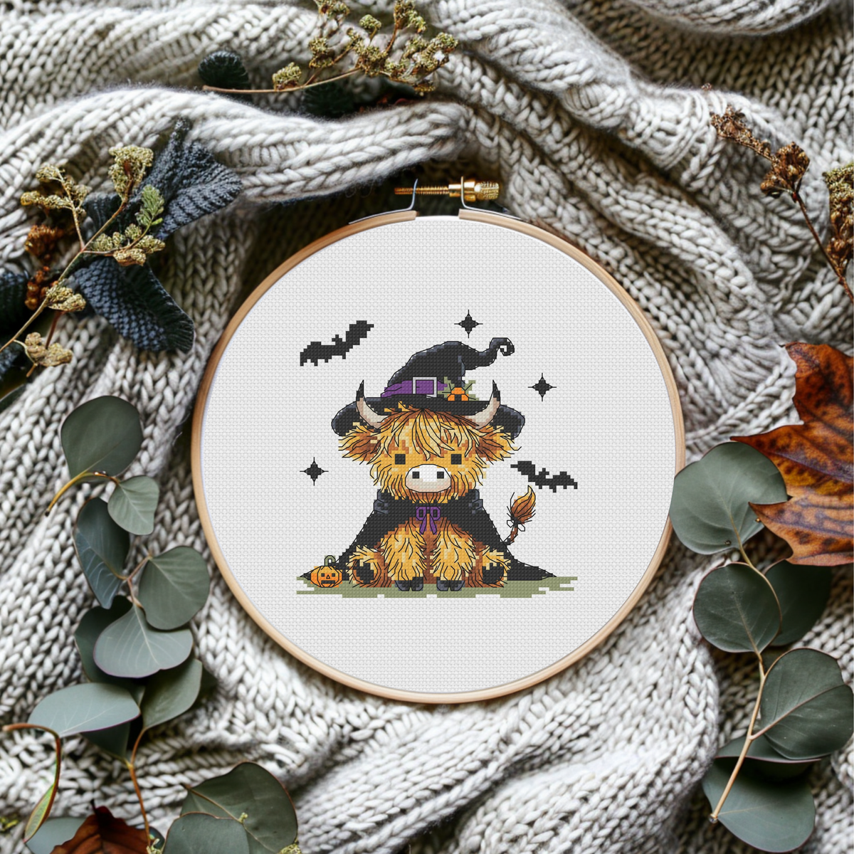 A circular embroidery hoop displaying a finished cross-stitch design of a cute black and yellow Scottish highland cow wearing a witch hat, styled on a cozy beige knit blanket with dried leaves and eucalyptus.