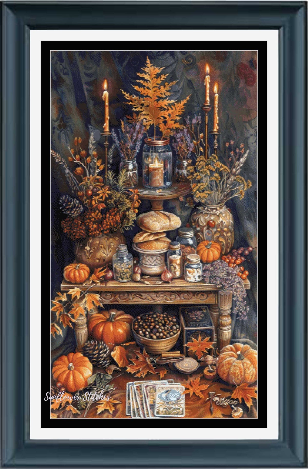 Framed still life cross stitch featuring an autumn tabletop scene with pumpkins, candles, flowers, bread, and fall leaves, presented in a dark teal frame with white matting.