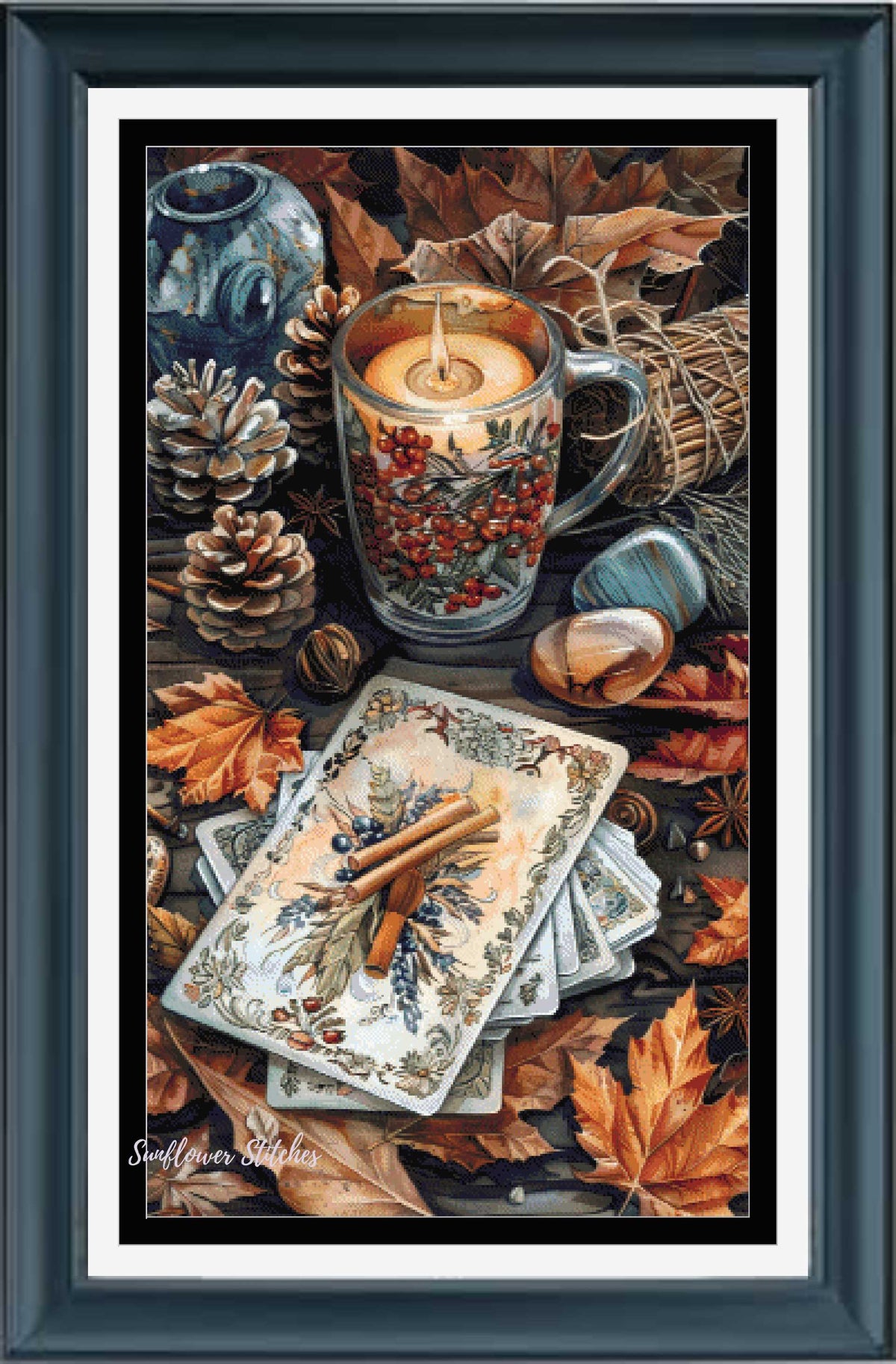 A framed still life artwork featuring a lit candle in a glass mug filled with red berries, surrounded by autumn leaves, pinecones, a blue vase, and a stack of illustrated cards on a dark tabletop. The piece is presented in a dark teal frame with a white mat.