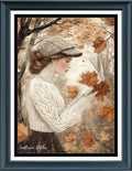 A framed cross stitch featuring an illustration of a woman in a cream cable-knit sweater and matching cap, holding autumn leaves in a tree-lined setting. The print is matted with a white border and enclosed in a dark teal-blue frame.