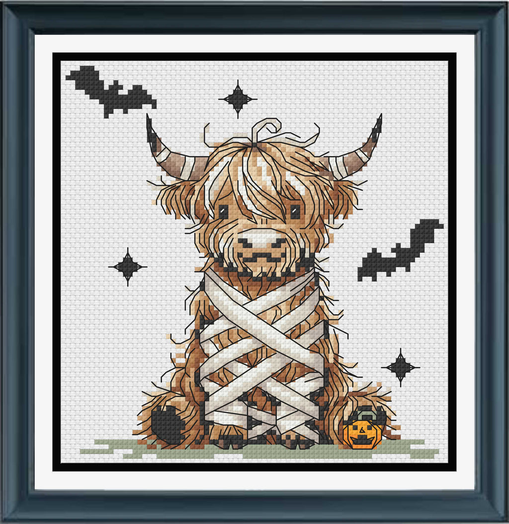 A framed cross-stitch design mock up featuring a fluffy Highland cow wrapped in white bandages, with black bats, starbursts, and a small orange pumpkin on a white fabric background. 