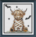 A framed cross-stitch design mock up featuring a fluffy Highland cow wrapped in white bandages, with black bats, starbursts, and a small orange pumpkin on a white fabric background. 