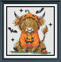 A framed cross-stitch of a fluffy Highland cow wearing an orange pumpkin costume, seated on a small patch of green grass with bats, stars, and a jack-o'-lantern around it. 