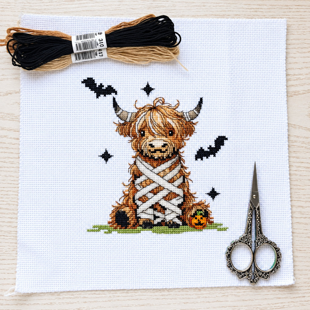 A completed cross-stitch design of a mummy Highland cow on white fabric, displayed with embroidery floss skeins, and decorative scissors, on a light wood background.