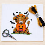 A completed cross-stitch design of a cute Highland cow in a pumpkin costume, displayed on a white fabric square alongside embroidery floss, scissors, and a thread spool on a light wood background.