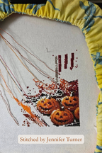 A close-up of a cross-stitch embroidery in progress on white fabric, showing stitched jack-o'-lantern pumpkins, orange leaves, and a candle with flames. The fabric is gathered at the top by a yellow and blue patterned cover, with the text 'Stitched by Jennifer Turner' at the bottom.