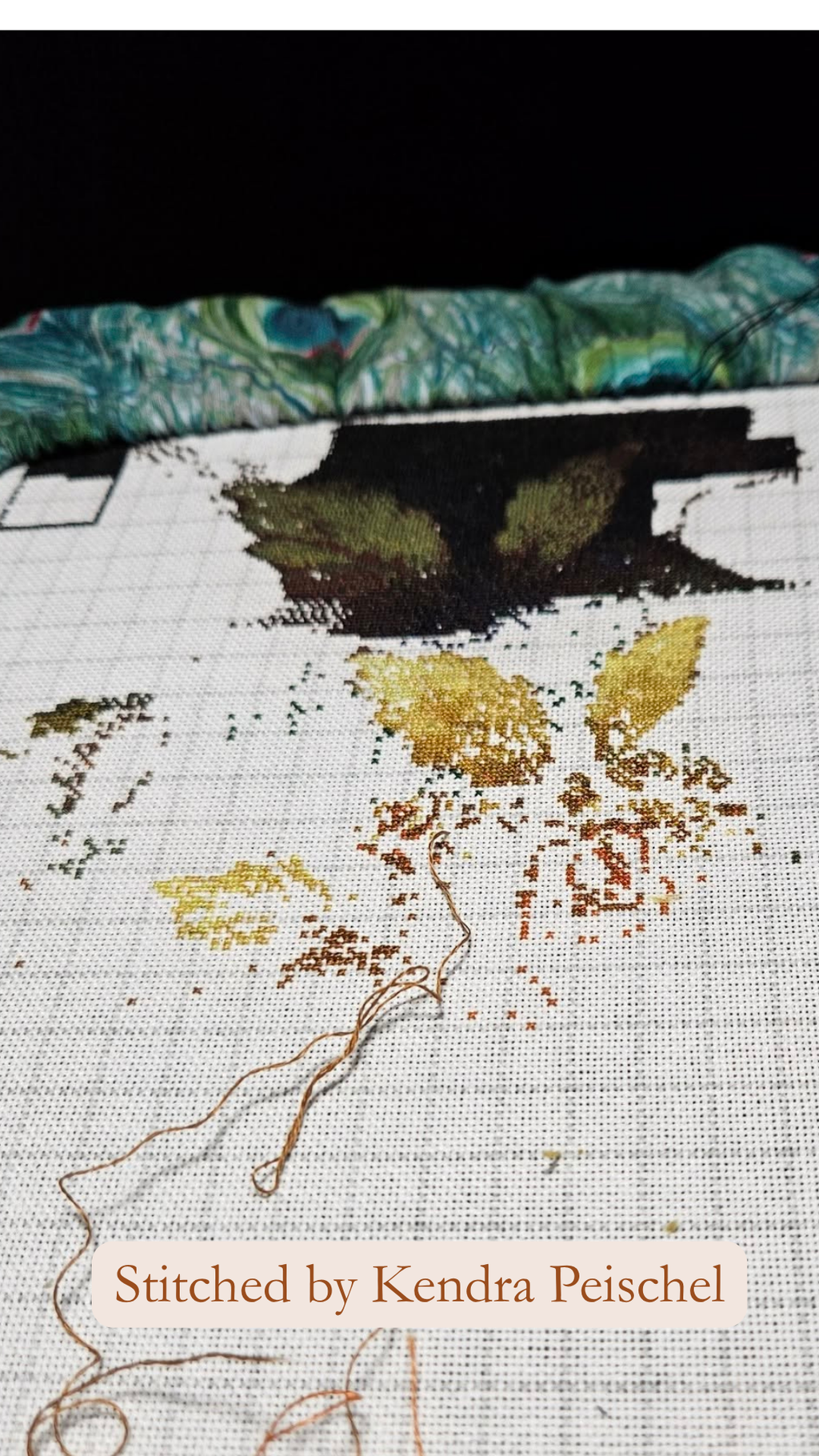 A close-up of a cross-stitch embroidery in progress on white Aida fabric, showing partial stitching with gold, brown, and black thread details. A teal and green patterned fabric roll is visible at the top, and a peach label at the bottom reads 'Stitched by Kendra Peischel'.