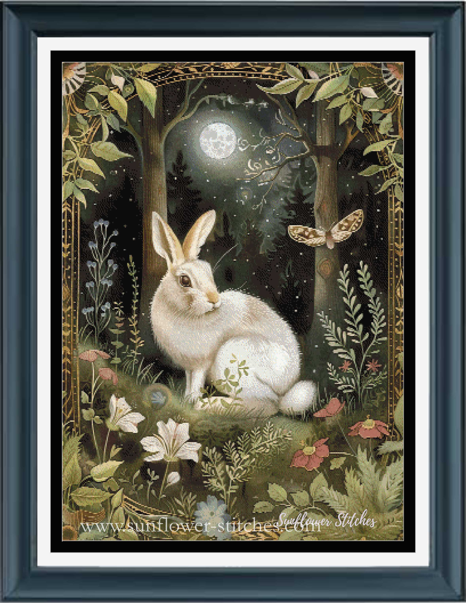 A framed cross stitch featuring a white rabbit sitting in a moonlit forest with flowers, foliage, a butterfly, and a decorative border. The artwork is presented in a dark blue frame with white matting.