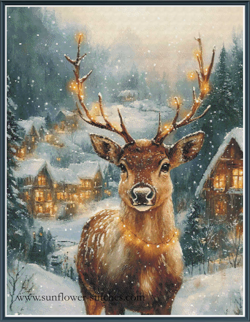 A festive winter landscape cross stitch featuring a brown stag with large antlers standing in the snow, surrounded by glowing holiday cottages and pine trees under falling snow.